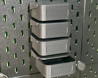 以下が含まれることがあります： ダークグレーのペグボードに取り付けられた、マットブラックの長方形収納ボックス4段重ね。各ボックスは滑らかな外観で、前面には長方形のくぼみがあります。一番下のボックスは少し開いており、内部が空洞になっているのが見えます。
