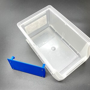 May include: A clear plastic storage container and a blue bracket. The container is rectangular with rounded corners and a slightly angled design. The blue bracket is rectangular with a small raised section on each end. The container is empty.