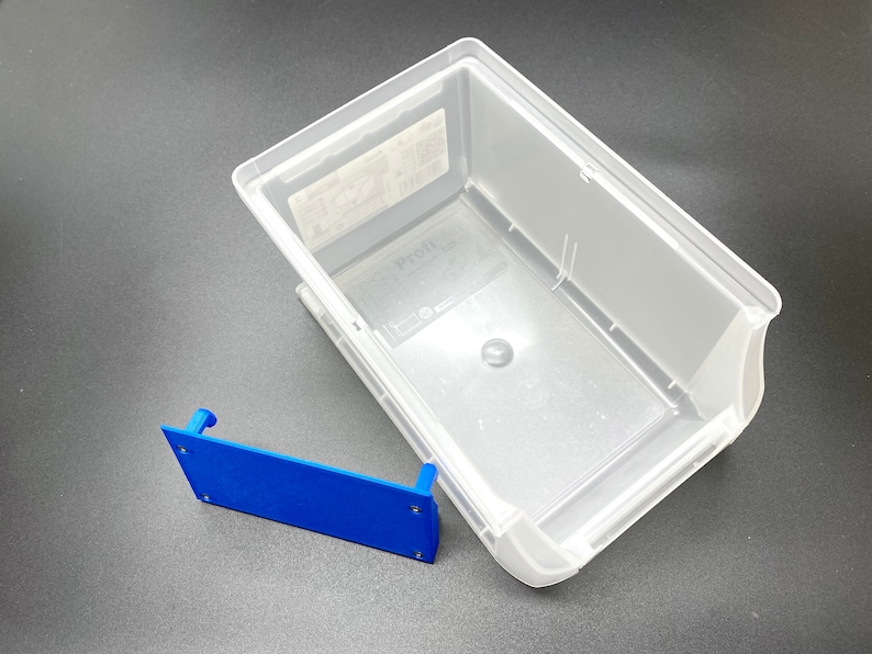 May include: A clear plastic storage container and a blue bracket. The container is rectangular with rounded corners and a slightly angled design. The blue bracket is rectangular with a small raised section on each end. The container is empty.