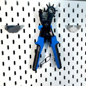 Könnte beinhalten: Ein schwarzes und blaues Stanzwerkzeug mit einem sternförmigen Stanzkopf, das an einer weißen Lochwand mit zwei grauen Kunststoff-Lochwandhaken hängt.