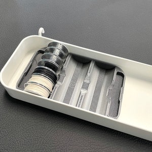 May include: A white plastic organizer with a built-in compartment holding spools of black, white, and silver thread. The organizer has a gray mesh insert and a hook for hanging.