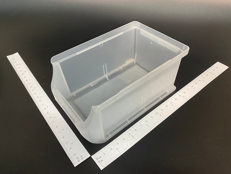 May include: A clear plastic storage container, rectangular with curved sides and a handle cutout. Two rulers are angled towards the bin. The container is empty, against a dark background. Measurements are in centimetres.