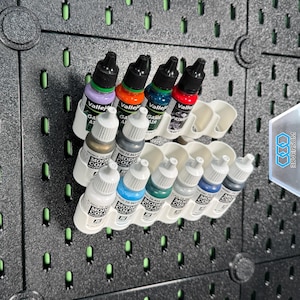 Könnte beinhalten: Eine Sammlung von Vallejo- und Model Color-Acrylfarben in einer weißen Halterung, montiert auf einer schwarzen Lochwand. Die Farben umfassen Lila, Orange, Grün, Rot, Weiß, Grau und Blau.