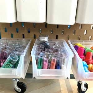 May include: A mobile craft storage unit with clear plastic bins. The bins are filled with colourful glitter, small plastic stamps, and other craft supplies. White containers hang above the bins, and the unit is set against a pegboard.