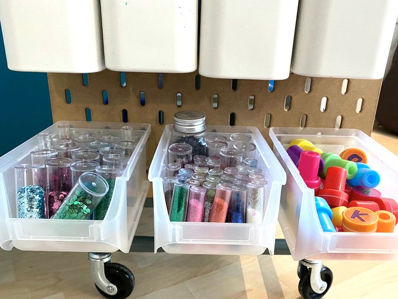 May include: A mobile craft storage unit with clear plastic bins. The bins are filled with colourful glitter, small plastic stamps, and other craft supplies. White containers hang above the bins, and the unit is set against a pegboard.