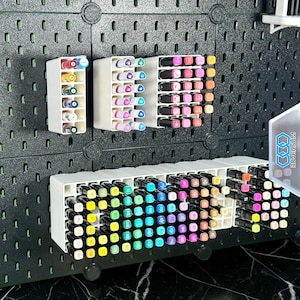 Könnte beinhalten: Weiße Kunststoff-Markerhalter mit verschiedenen farbigen Markern sind an einer schwarzen Lochwand befestigt. Die Marker sind in Reihen angeordnet und zeigen eine große Auswahl an Farben. Die Halter sind so konzipiert, dass die Marker organisiert und zugänglich sind.