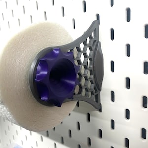 K&ouml;nnte beinhalten: Ein grauer und violetter Papierhandtuchhalter, der an einer wei&szlig;en Lochwand befestigt ist. Der Halter hat ein geometrisches Design und einen violetten, sternf&ouml;rmigen Knopf. Eine Rolle wei&szlig;er Papierhandt&uuml;cher befindet sich im Halter.
