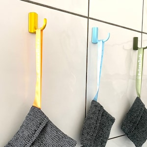 Könnte beinhalten: Drei Haken mit Stoffschlaufen, die daran hängen. Die Haken sind gelb, blau und grün. Die Stoffschlaufen sind grau und haben den Text "HomeBody" aufgedruckt.