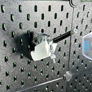 May include: An airbrush mounted on a black pegboard. The airbrush is silver and black, with a white holder. The pegboard has a grid of holes with green accents. A clear plastic piece with the text "3DVERSUM" is in the background.