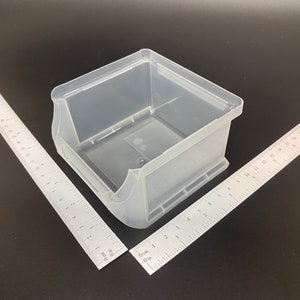 May include: A clear, translucent plastic storage bin with a rectangular shape and curved sides. Two metal rulers are positioned at angles to the bin. The rulers are marked with centimetre measurements.