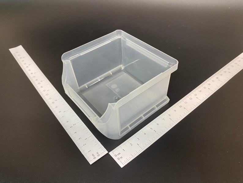 May include: A clear, translucent plastic storage bin with a rectangular shape and curved sides. Two metal rulers are positioned at angles to the bin. The rulers are marked with centimetre measurements.