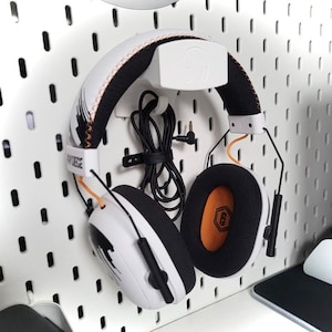 Op de afbeelding: Witte en zwarte gaming-hoofdtelefoon met oranje accenten, gemonteerd op een witte pegboard. De hoofdtelefoon heeft een zwart snoer en een logo op de oorschelpen. De oorschelpen zijn zwart met oranje vulling.