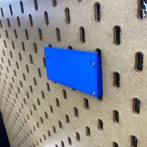 May include: A vibrant blue rectangular item is attached to a pegboard. The pegboard is made of light brown wood and features a grid of oval holes. The blue object is fixed with small silver screws.