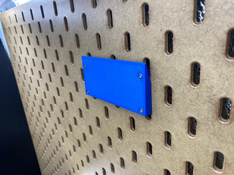 May include: A vibrant blue rectangular item is attached to a pegboard. The pegboard is made of light brown wood and features a grid of oval holes. The blue object is fixed with small silver screws.