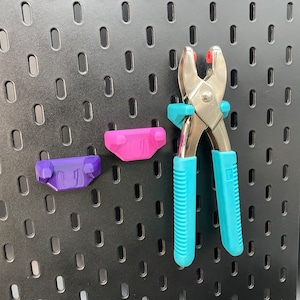 Può includere: Uno strumento in metallo argentato con manici in plastica blu e rosa è appeso a una bacheca nera. Lo strumento viene utilizzato per fissare gli occhielli al tessuto.