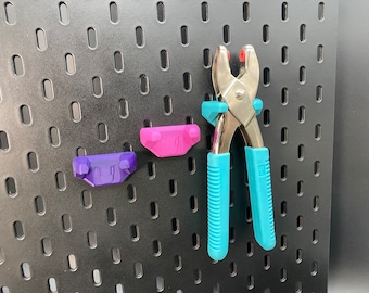 以下が含まれることがあります： 青いプラスチック製のハンドルとピンクのプラスチック製のハンドルが付いた銀色の金属製の工具が、黒いペグボードに掛けられています。この工具は、生地にアイレットを取り付けるために使用されます。