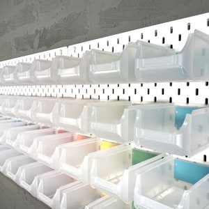 Op de afbeelding: Een wit pegboard met meerdere rijen doorzichtige plastic opbergbakken. De bakken zijn op het bord gemonteerd en hebben een licht matte uitstraling. De achtergrond is een grijze muur met textuur.
