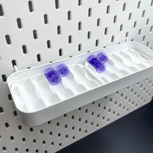 May include: A white pegboard with a white storage shelf and several purple suction cup accessories. The shelf is rectangular with divided compartments. The pegboard has evenly spaced holes.
