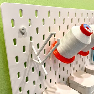 Könnte beinhalten: Weiße Lochwand mit verschiedenen Nähutensilien. Weiße Garnrollen mit roten und blauen Etiketten werden von grauen Halterungen gehalten. Zusätzliche weiße Aufbewahrungsbehälter sind sichtbar, zusammen mit einem grauen Stift und einer grünen Wand im Hintergrund.