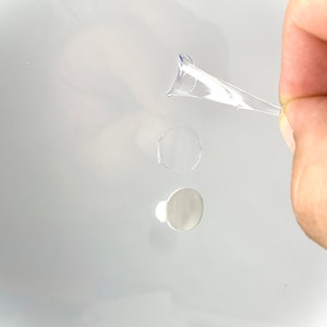 Peut inclure: Un autocollant adh&eacute;sif en plastique transparent de forme ronde. L'autocollant est partiellement d&eacute;coll&eacute; et est tenu par une main.