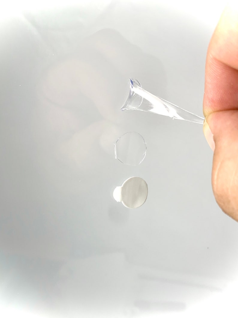 Peut inclure: Un autocollant adh&eacute;sif en plastique transparent de forme ronde. L'autocollant est partiellement d&eacute;coll&eacute; et est tenu par une main.