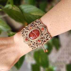 May include: A brown macrame bracelet with a large red agate stone in the center. The bracelet is decorated with smaller red agate stones around the center stone.