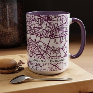 May include: A white ceramic mug with a purple map of London, England printed on the exterior. The mug features a purple interior and a matching purple handle. The text LONDON • ENGLAND is printed in black on the lower portion of the mug.
