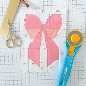 Op de afbeelding: Een roze stoffen strik op een witte achtergrond met bloemenprint. De strik is gemaakt van meerdere stofdelen die aan elkaar zijn genaaid. De stof wordt gesneden op een blauwe snijmat met een rolmes.