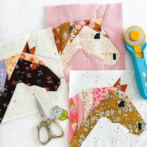 May include: A collection of colorful fabric horse head quilt blocks. The blocks feature various floral and patterned fabrics in shades of pink, brown, and white. A blue rotary cutter and gold scissors are also present.