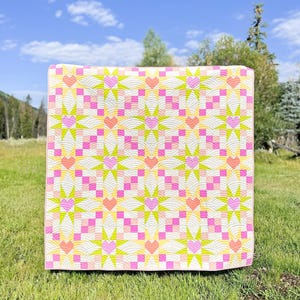 May include: A colorful quilt with a repeating pattern of pink hearts, yellow starbursts, and pink and yellow squares. The quilt is displayed outdoors on a grassy surface, with a blue sky and trees in the background. The quilt is made of soft fabric.