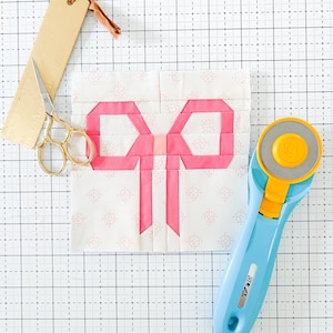 May include: A pink fabric bow on a white background with a pink polka dot pattern. The bow is made of fabric squares sewn together. The fabric is on a white cutting mat with a grid pattern. A rotary cutter and a pair of scissors are also visible.