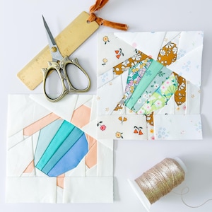 May include: An overhead shot of quilting supplies, including two fabric blocks with geometric patterns in white, peach, blue, and floral prints. A pair of gold scissors, a wooden ruler, and a spool of gold thread are also present.