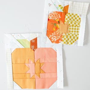 May include: Two colourful quilt blocks with pumpkin designs. One block features orange floral fabric, yellow gingham, and a star. The other block has peach and orange tones with a star design. Both blocks are bordered with white fabric.