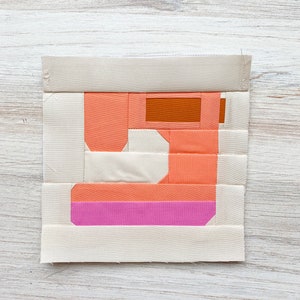 Op de afbeelding: Een patchwork quiltblok in de vorm van een naaimachine. Het blok is gemaakt van oranje, wit en roze stof.