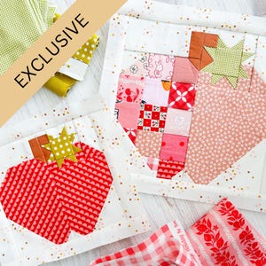 Pode incluir: Dois blocos de colcha inacabados com um design de morango em forma de coração. Os blocos são feitos com uma variedade de tecidos rosa, vermelho e branco. Os blocos são colocados sobre um fundo branco com glitter dourado.
