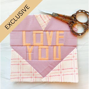 Könnte beinhalten: Ein rosa-weißer Patchwork-Quiltblock mit den Worten "I LOVE YOU" in Herzform. Der Block ist aus verschiedenen Stoffen gefertigt, darunter rosa und weißes Gingham.