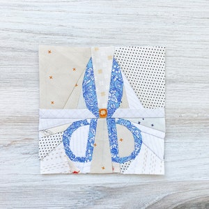 May include: A fabric art piece featuring a pair of scissors. The scissors are blue with a floral pattern and a gold-colored center. The background is a patchwork of various fabrics in neutral tones and patterns, including dots and crosses.