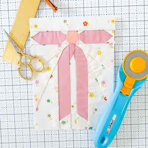 Può includere: Un fiocco di stoffa rosa e bianco con un motivo floreale. Il fiocco è su un tappetino da taglio bianco con un righello e un taglierino rotativo.