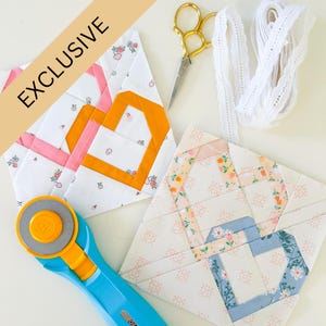 May include: Two heart-shaped quilt blocks with floral patterns and colorful accents, alongside gold scissors, white lace ribbon, and a blue rotary cutter. The word "EXCLUSIVE" is printed on a diagonal banner.