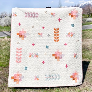 May include: A white quilt with a pink and orange floral pattern. The quilt is made up of different blocks with various designs, including flowers, leaves, and geometric shapes. The quilt is displayed on a grassy field.