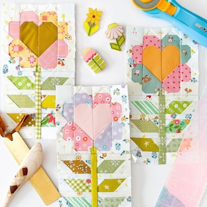 May include: Three fabric flower quilt blocks with heart-shaped centers in pink, yellow, and orange. The blocks are surrounded by quilting tools and flower-shaped pins. The blocks are made of various patterned fabrics.