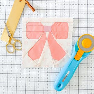 May include: A pink fabric bow with a white background, a gold ruler, a pair of gold scissors, and a blue rotary cutter on a white grid cutting mat.