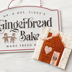 May include: A small gingerbread house quilt block made with brown and white patterned fabric. The house features a pink heart and a nutcracker figure in the window. The quilt block is on a white surface next to a sign that reads "Mr. & Mrs. Claus's Gingerbread Bake Shop Made Fresh Daily".