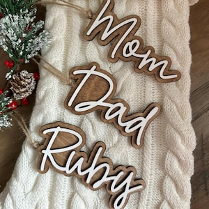 May include: Three wooden Christmas stocking ornaments with white lettering. The ornaments say "Mom", "Dad", and "Ruby".