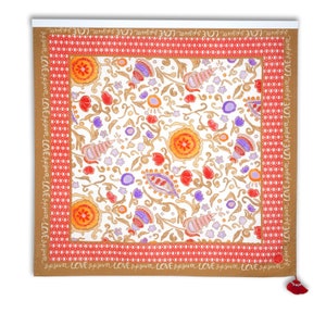 Puede incluir: Textil cuadrado con fondo blanco y un colorido estampado floral. El diseño incluye flores naranjas, rojas y moradas, junto con vides doradas y formas de conchas marinas. El borde es marrón con la palabra "LOVE" repetida. Una borla roja cuelga en la parte inferior.