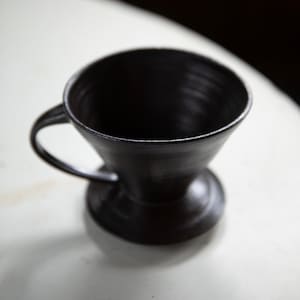 May include: A black ceramic coffee filter with a handle and a wide base. The filter is designed for pour-over coffee brewing.