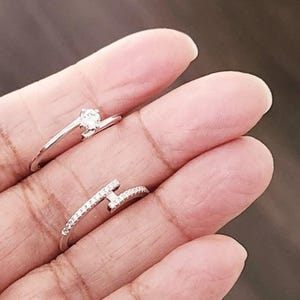 May include: Two silver rings are shown. One ring has a single, round, clear gemstone. The other ring features a nail-head design, with small, clear gemstones along the band and nail head.