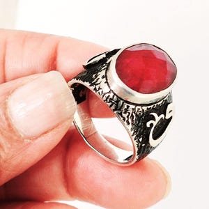May include: A silver ring featuring a large, oval-cut, red gemstone. The ring has a textured, dark band with decorative patterns. The gemstone is set in a silver bezel, creating a striking contrast.