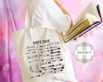May include: Cream-colored tote bag with a black "DATE DUE" library card design. The bag features a list of dates and the Dixie Design Co. logo. The bag is held by a person wearing glasses and a white shirt, holding a book.