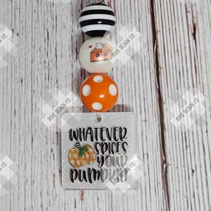 May include: A fall-themed keychain with a silver clasp. It features three beads: a black and white striped bead, a white bead with a pumpkin pie graphic, and an orange polka dot bead. A square charm reads "WHATEVER SPICES YOUR PUMPKIN".